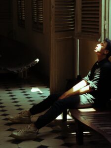 A young man sits in a dimly lit room, soaked in sunlight, depicting introspection.